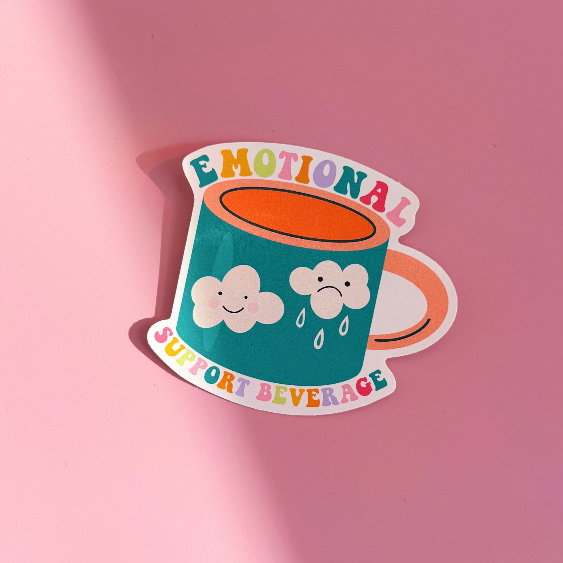 A waterproof sticker, featuring an illustration of a mug with a sad and a happy cloud on the front. Around the mug are the words 'emotional support beverage'.