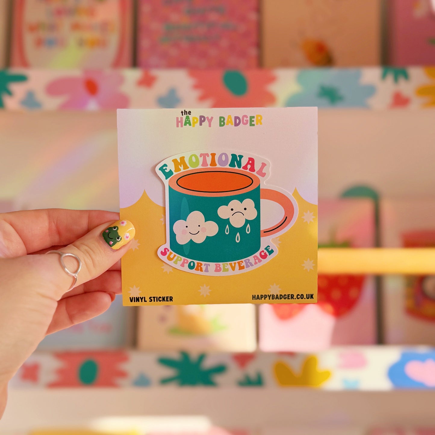 A waterproof sticker, featuring an illustration of a mug with a sad and a happy cloud on the front. Around the mug are the words 'emotional support beverage'.