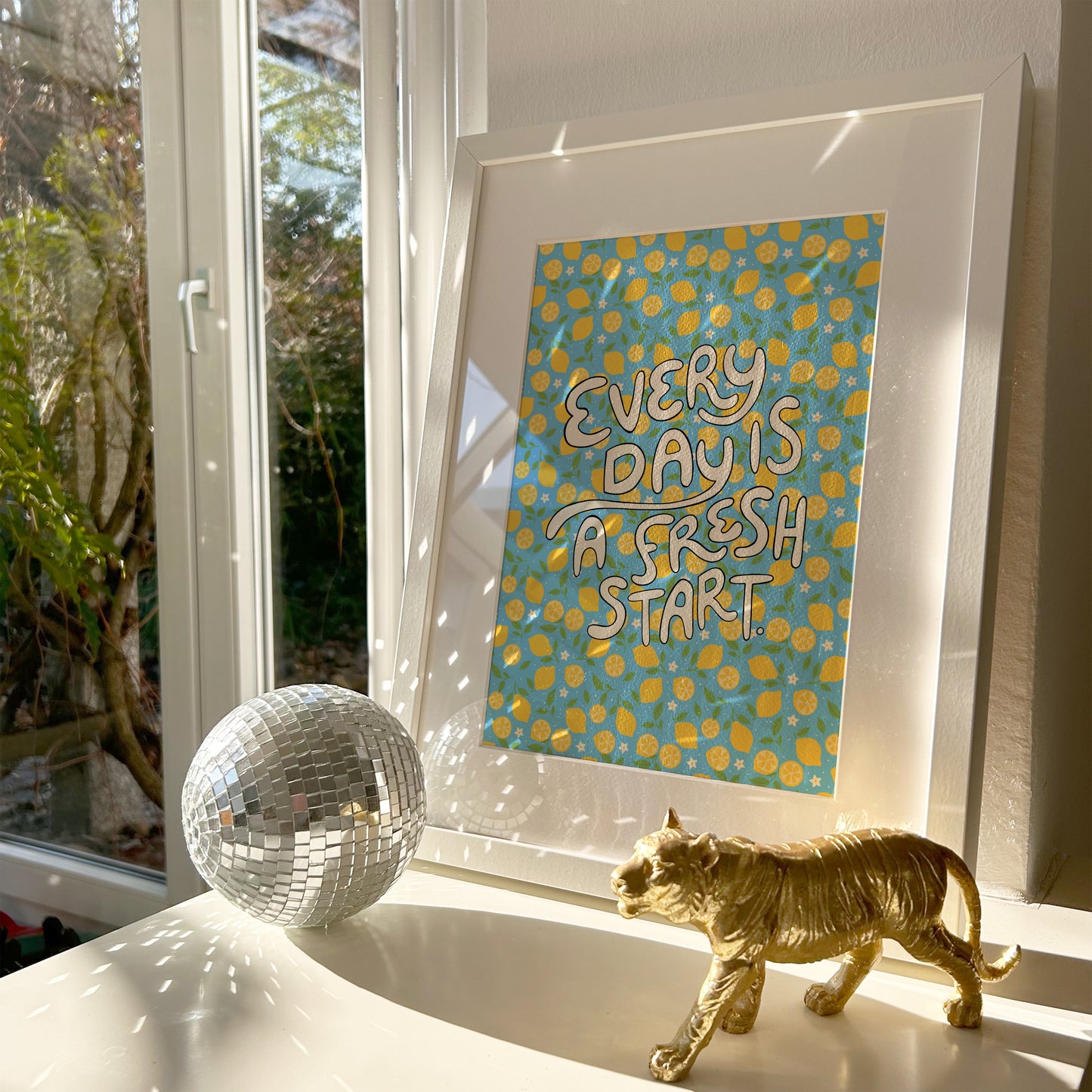 A bright quote print, with hand lettered words reading 'every day is a fresh start' on a blue background with a lemon pattern.