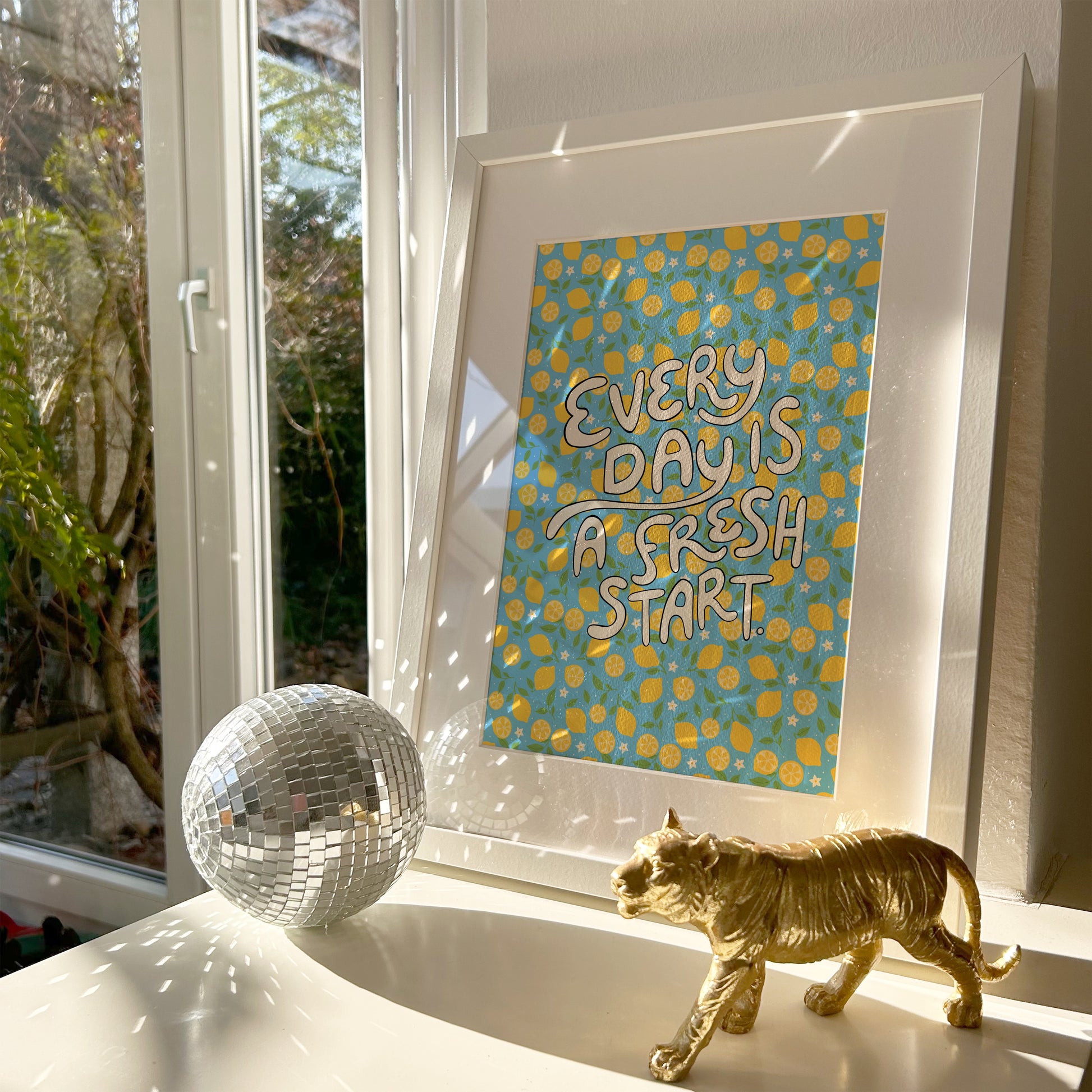 A bright quote print, with hand lettered words reading 'every day is a fresh start' on a blue background with a lemon pattern.