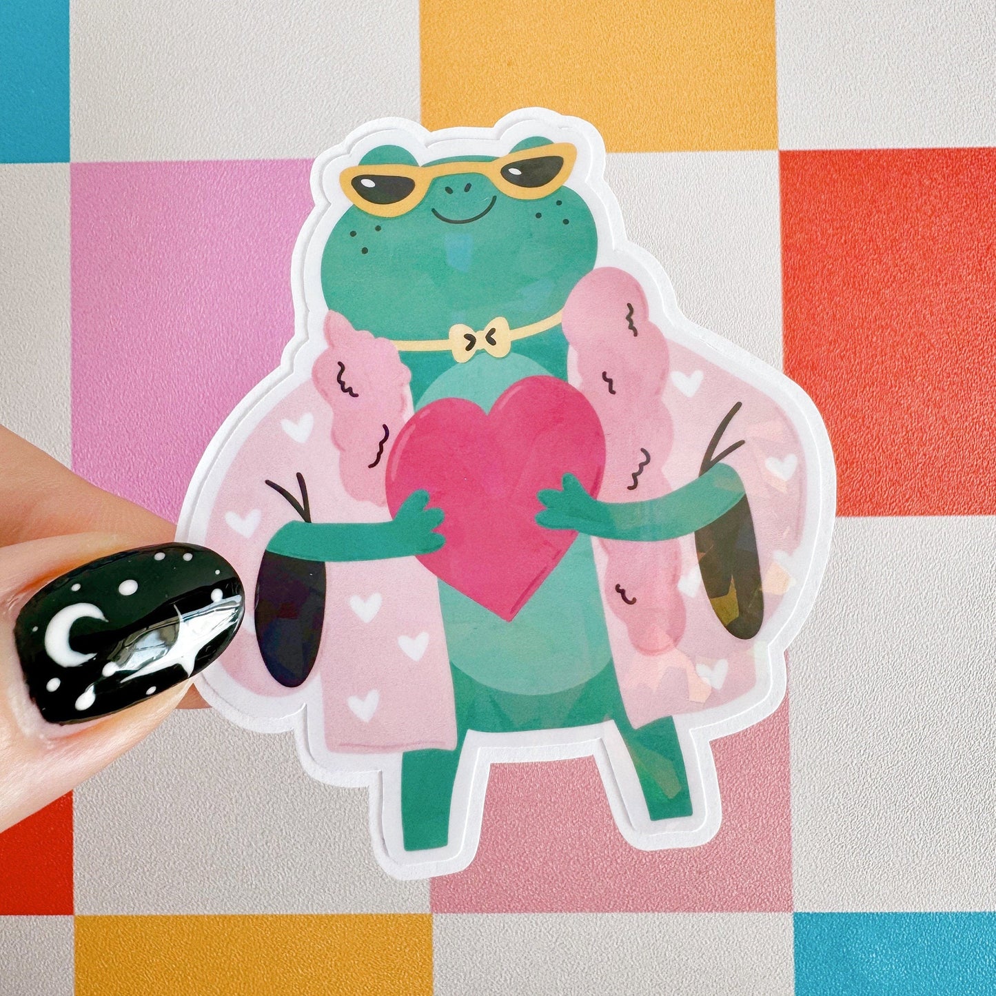 A holographic sticker, of a green frog with a pink coat on holding a love heart. The sticker has an easy peel lip.