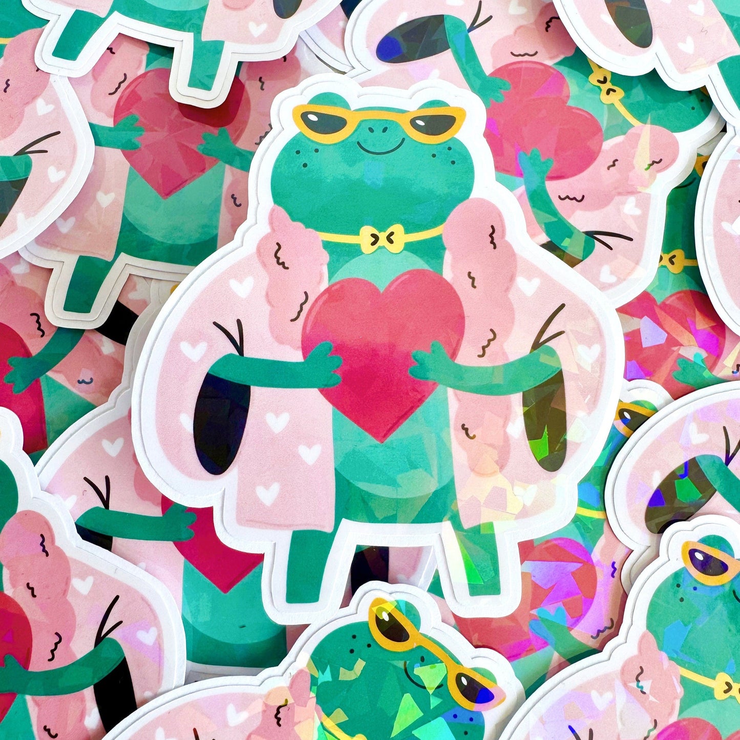 A holographic sticker, of a green frog with a pink coat on holding a love heart. The sticker has an easy peel lip.