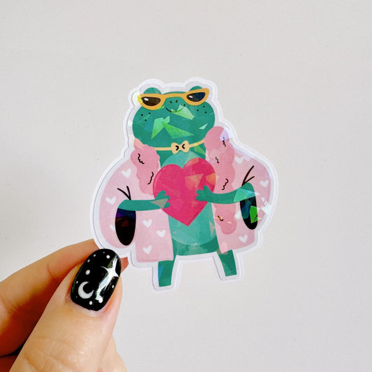 A holographic sticker, of a green frog with a pink coat on holding a love heart. The sticker has an easy peel lip.