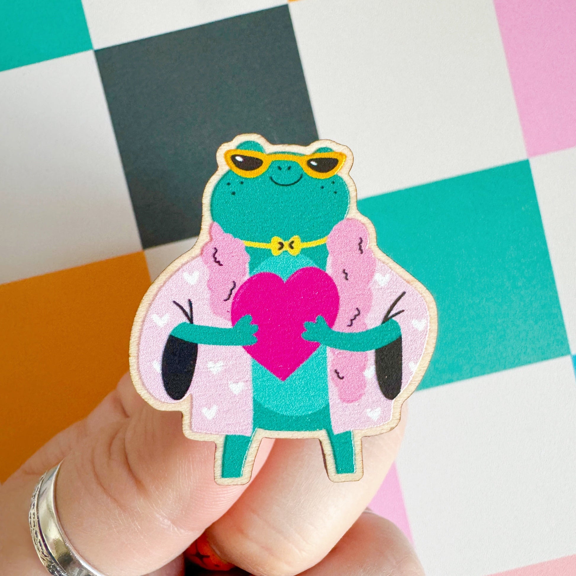 A cute illustration of a frog in sunglasses and a pink coat, holding a love heart, on an eco friendly wooden pin badge with two back fastenings.