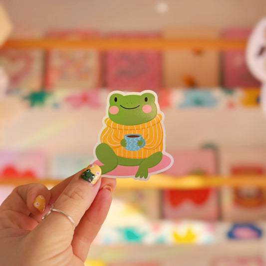 Hand holding a sticker of a frog wearing a sweater with a cup in front of a colourful background.