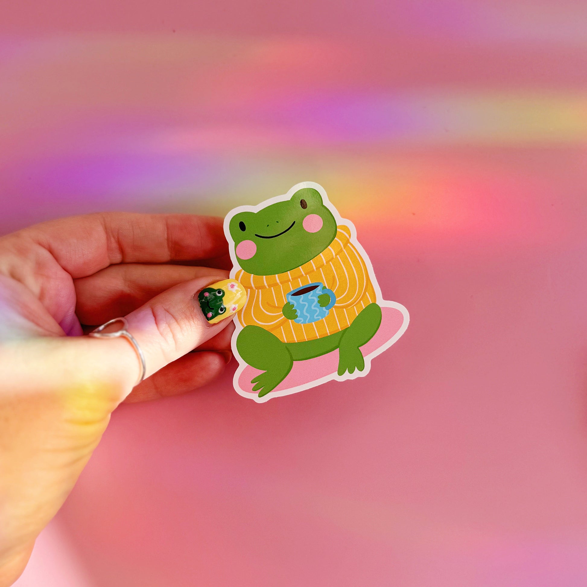 Hand holding a sticker of a frog wearing a sweater with a cup in front of a colourful background.