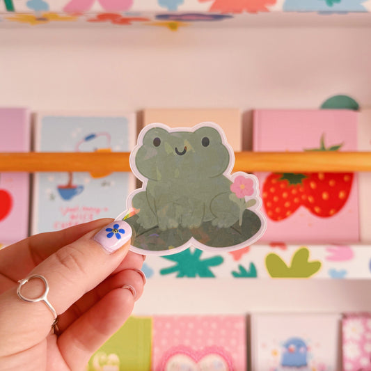 A holographic sticker featuring a frog sat on a lily pad.