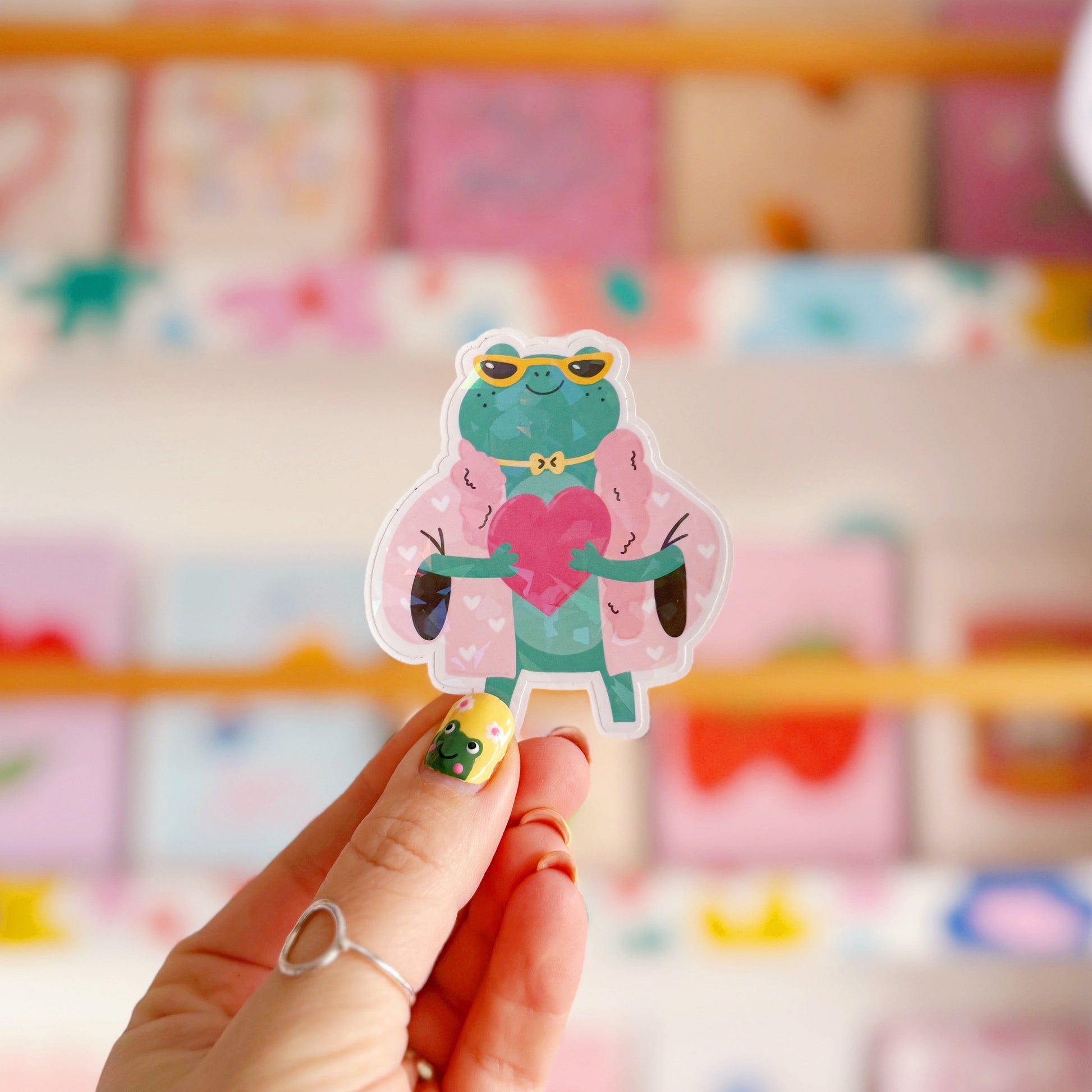 A holographic sticker, of a green frog with a pink coat on holding a love heart. The sticker has an easy peel lip.
