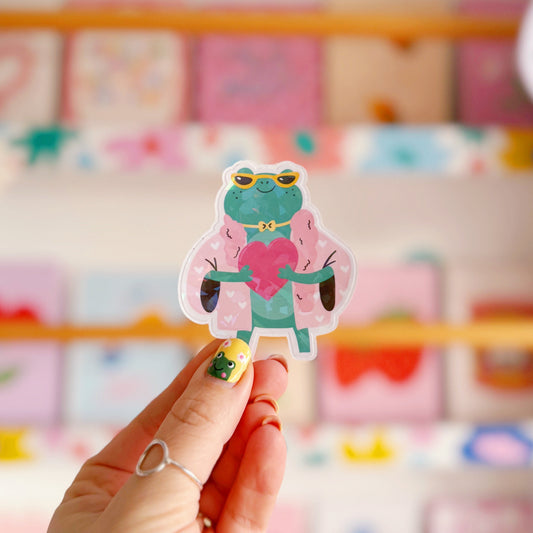 A holographic sticker, of a green frog with a pink coat on holding a love heart. The sticker has an easy peel lip.