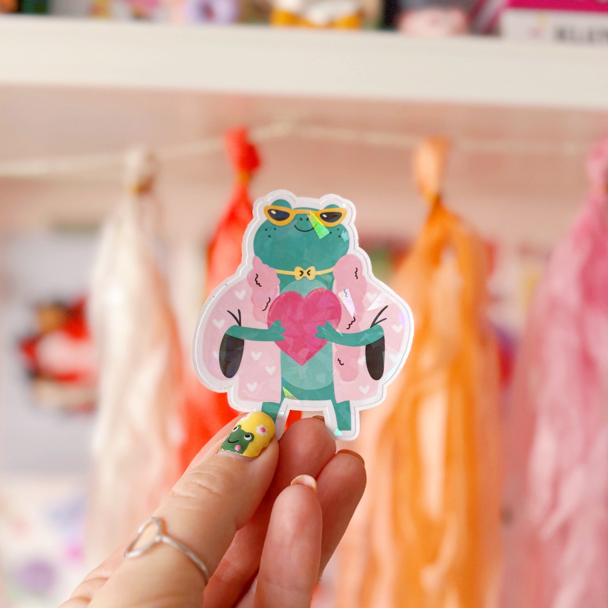 A holographic sticker, of a green frog with a pink coat on holding a love heart. The sticker has an easy peel lip.