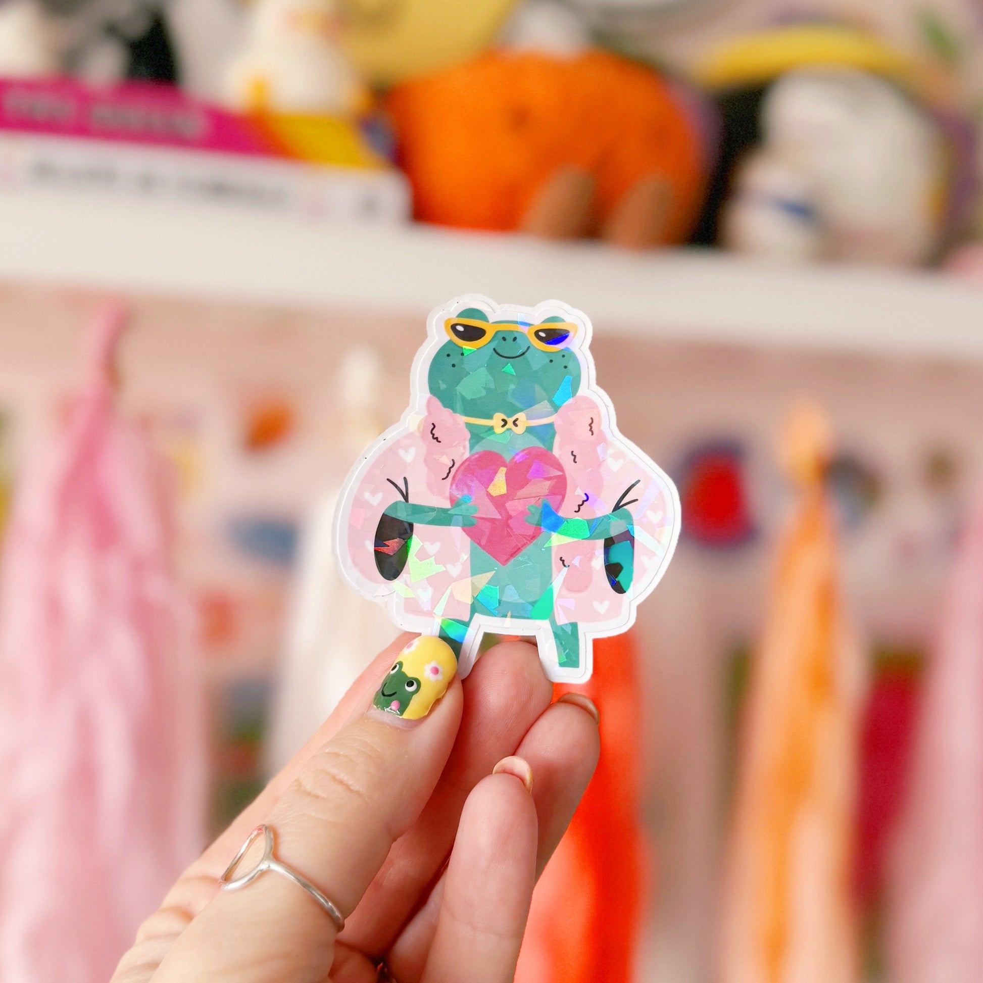 A holographic sticker, of a green frog with a pink coat on holding a love heart. The sticker has an easy peel lip.