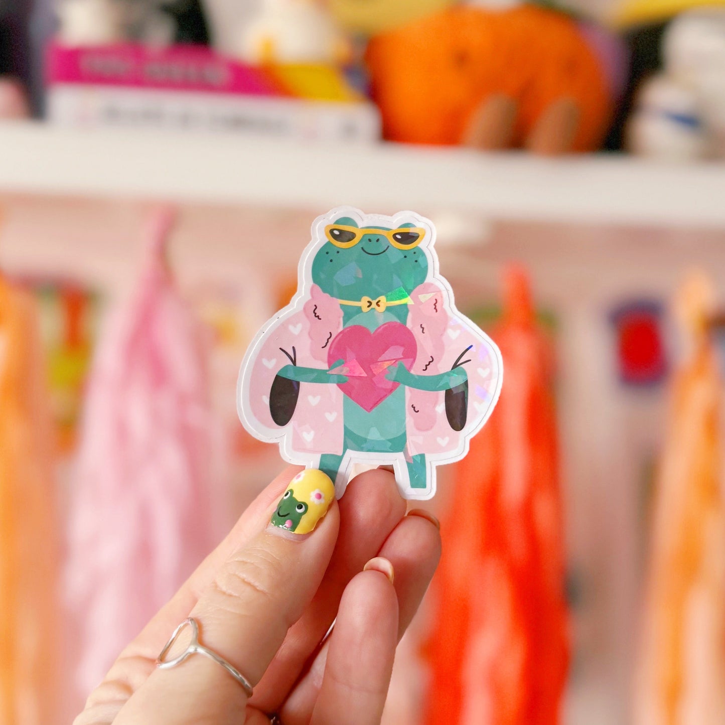 A holographic sticker, of a green frog with a pink coat on holding a love heart. The sticker has an easy peel lip.