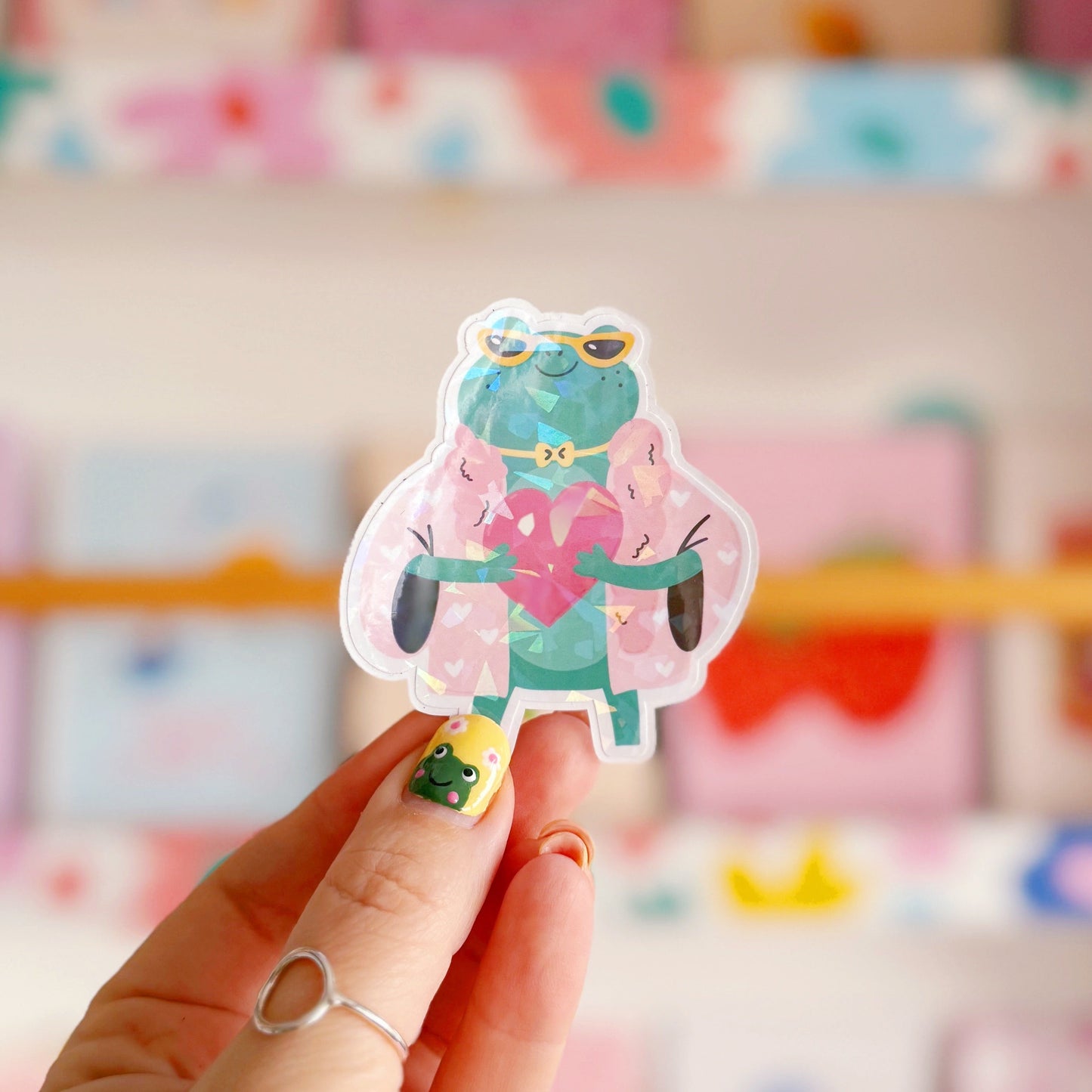 A holographic sticker, of a green frog with a pink coat on holding a love heart. The sticker has an easy peel lip.