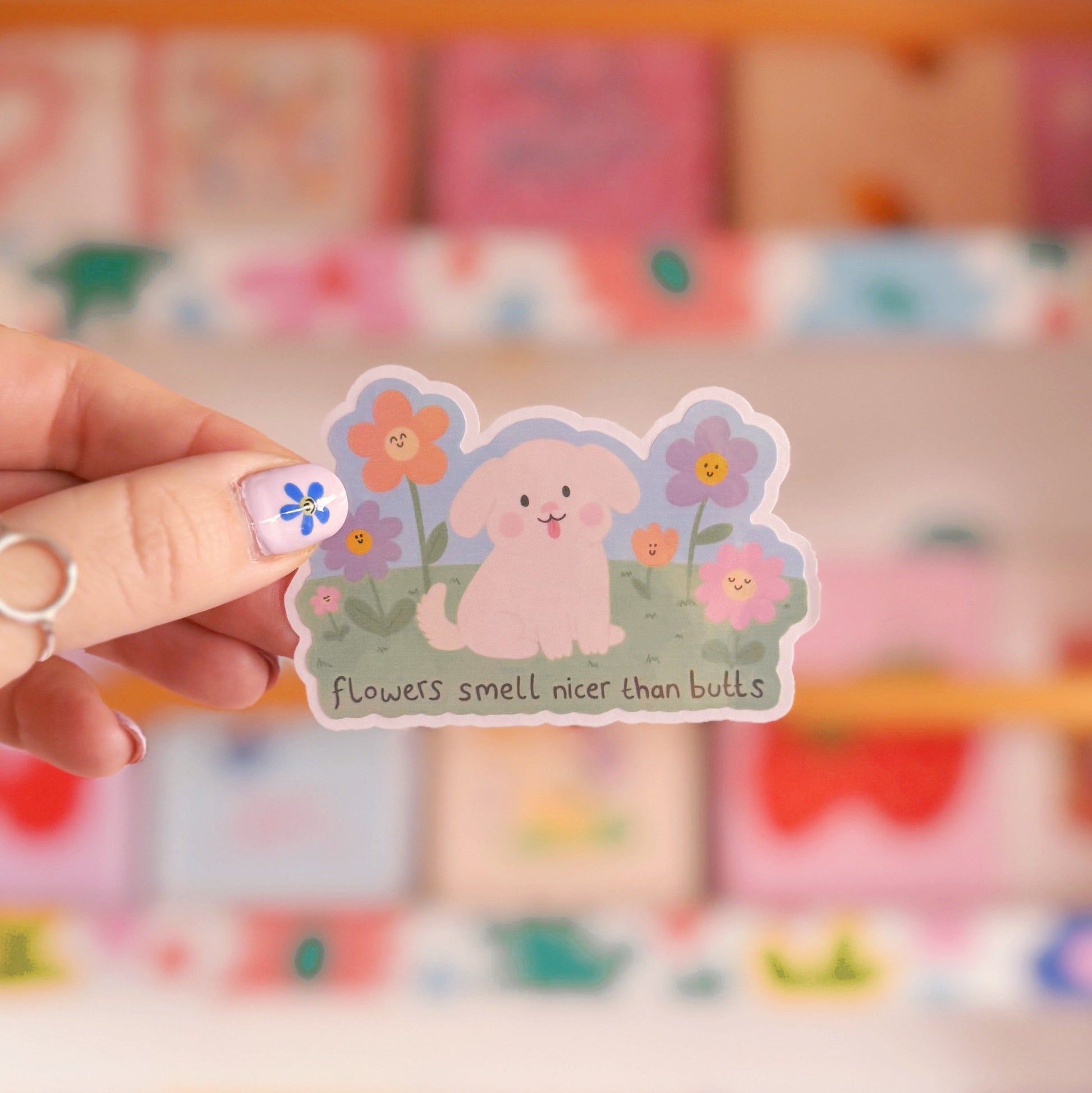 An illustrated sticker of a dog sat amongst flowers, with the quote 'flowers smell nicer than butts' underneath'.