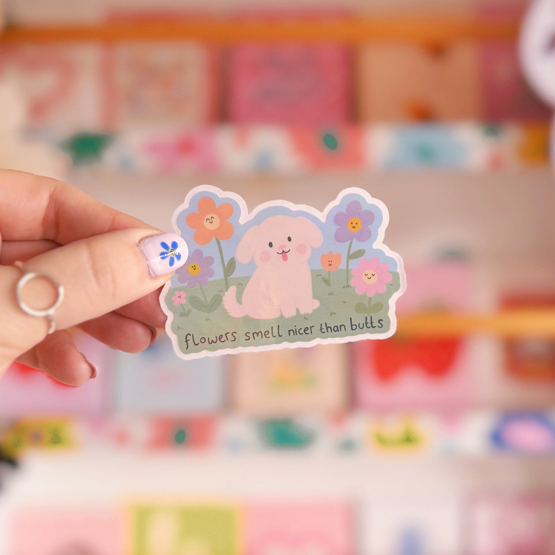 An illustrated sticker of a dog sat amongst flowers, with the quote 'flowers smell nicer than butts' underneath'.