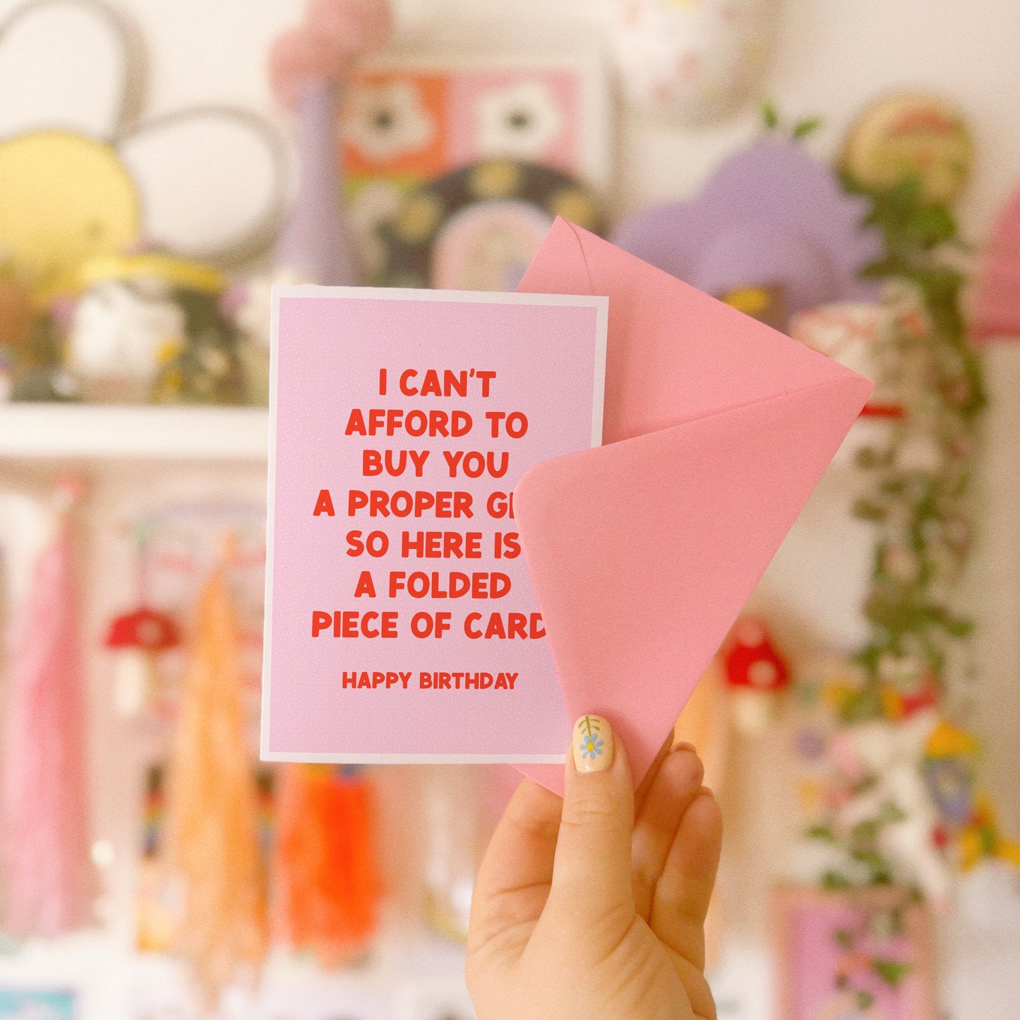 Hand holding a pink card with red text and a pink envelope against a colorful, blurred background.