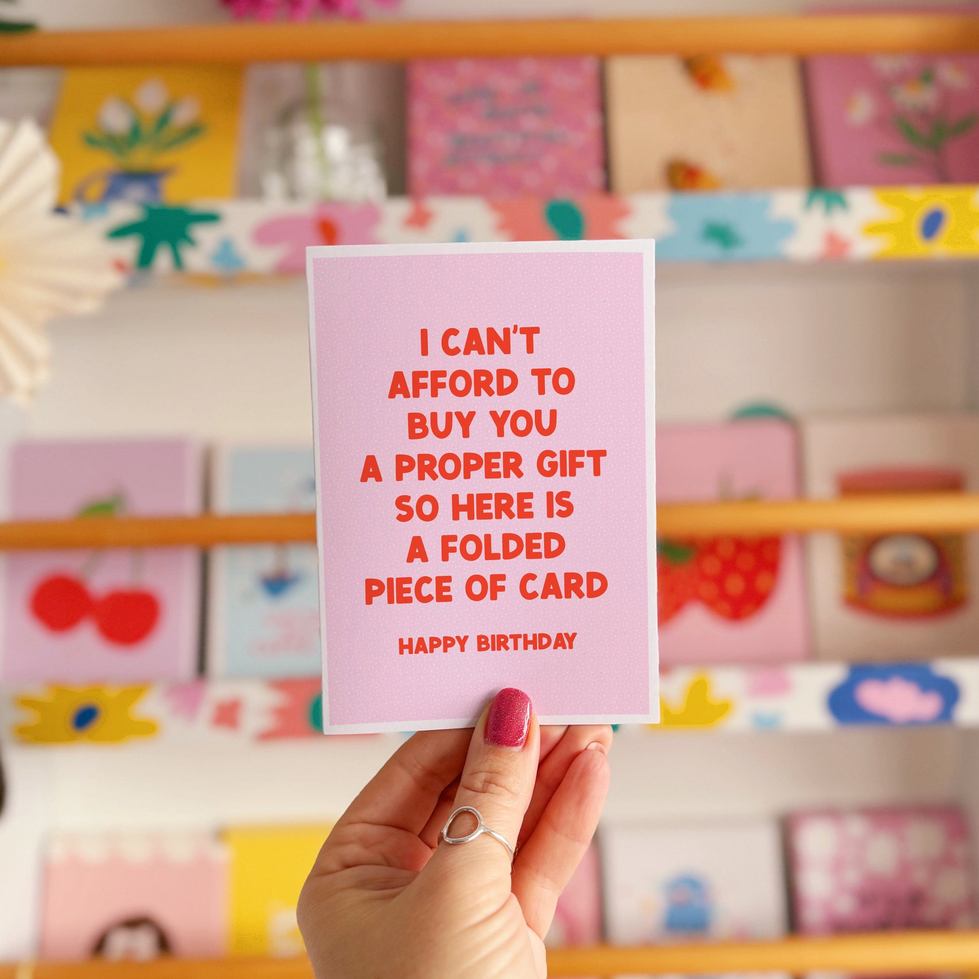 Hand holding a pink card with red text in front of a display of colorful greeting cards.