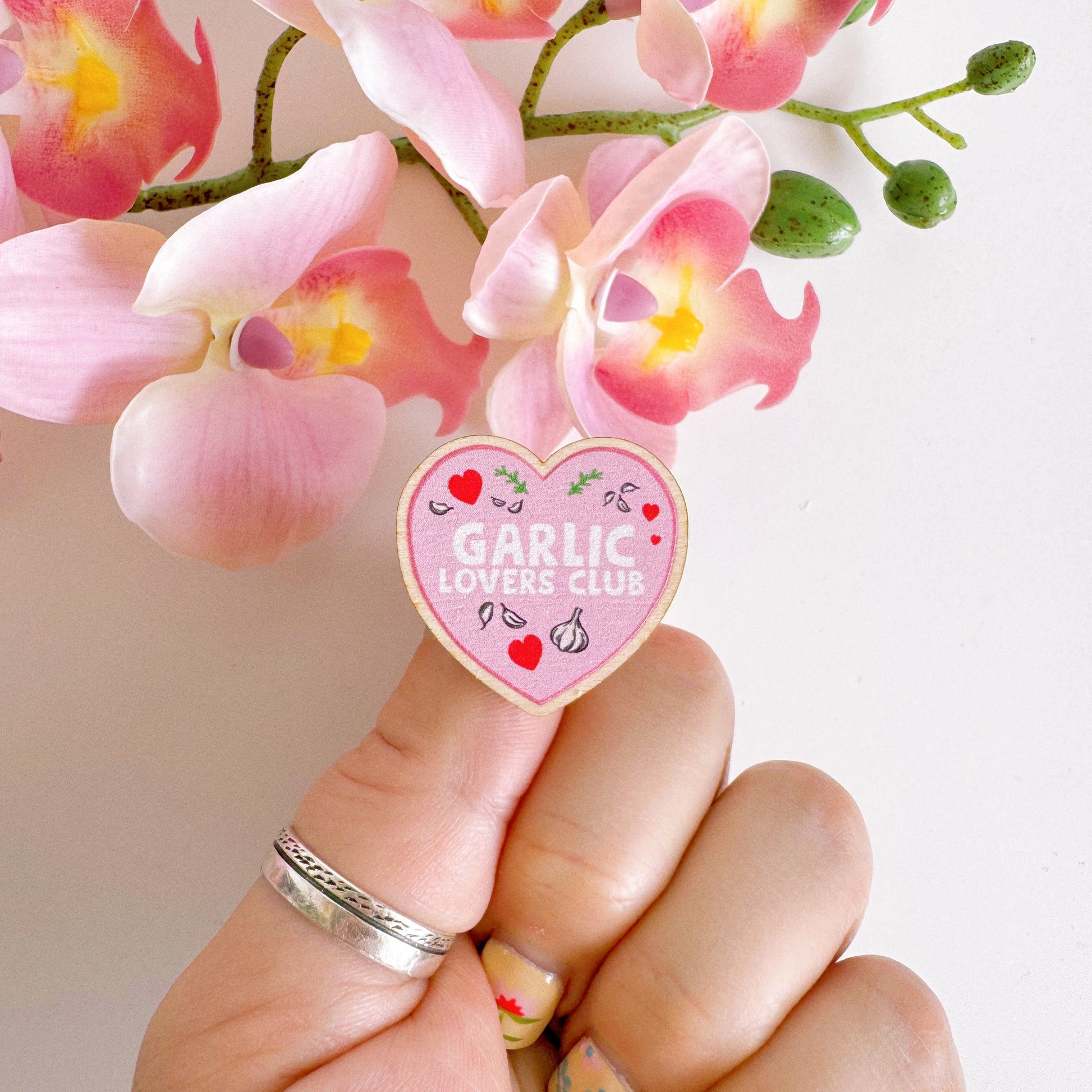 A pink heart shaped wooden pin badge, with the words 'garlic lovers club' on the front