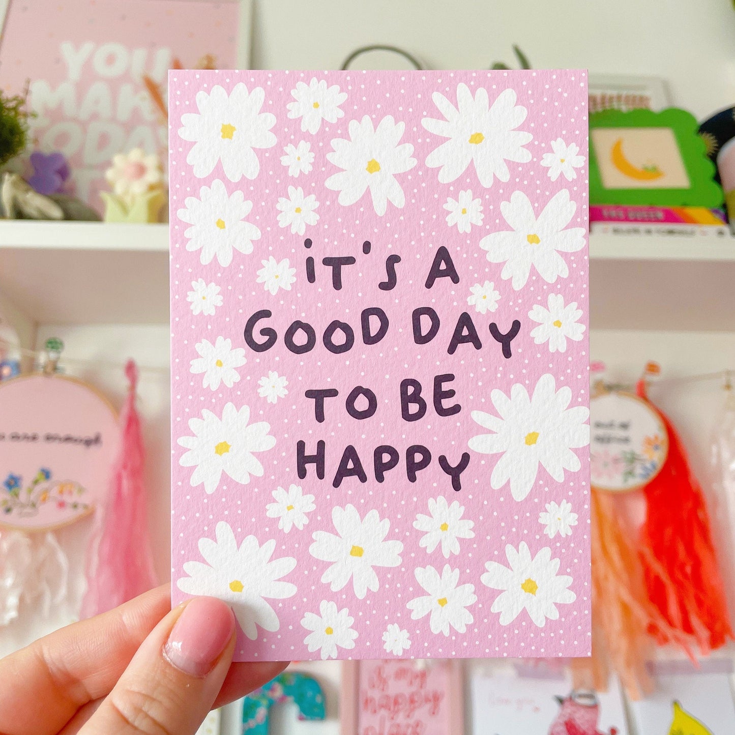 A textured postcard, with the words it's a good day to be happy, on the front, on a pink background with white daises.