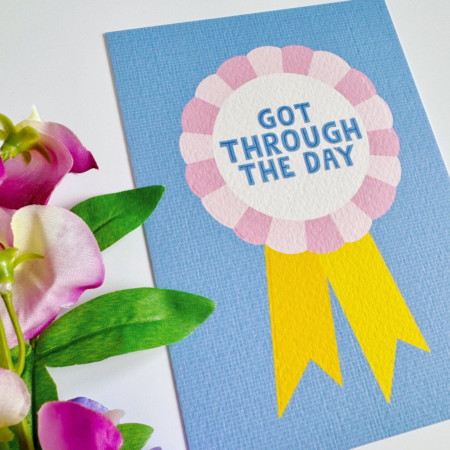 A blue postcard, with a rosette in pink and yellow, saying got through the day.