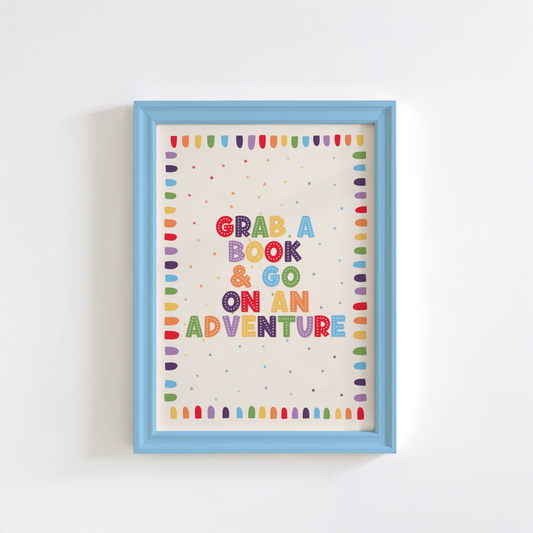 A framed children's book-themed art print. The print features the phrase 'GRAB A BOOK & GO ON AN ADVENTURE' in colourful letters with a rainbow border.