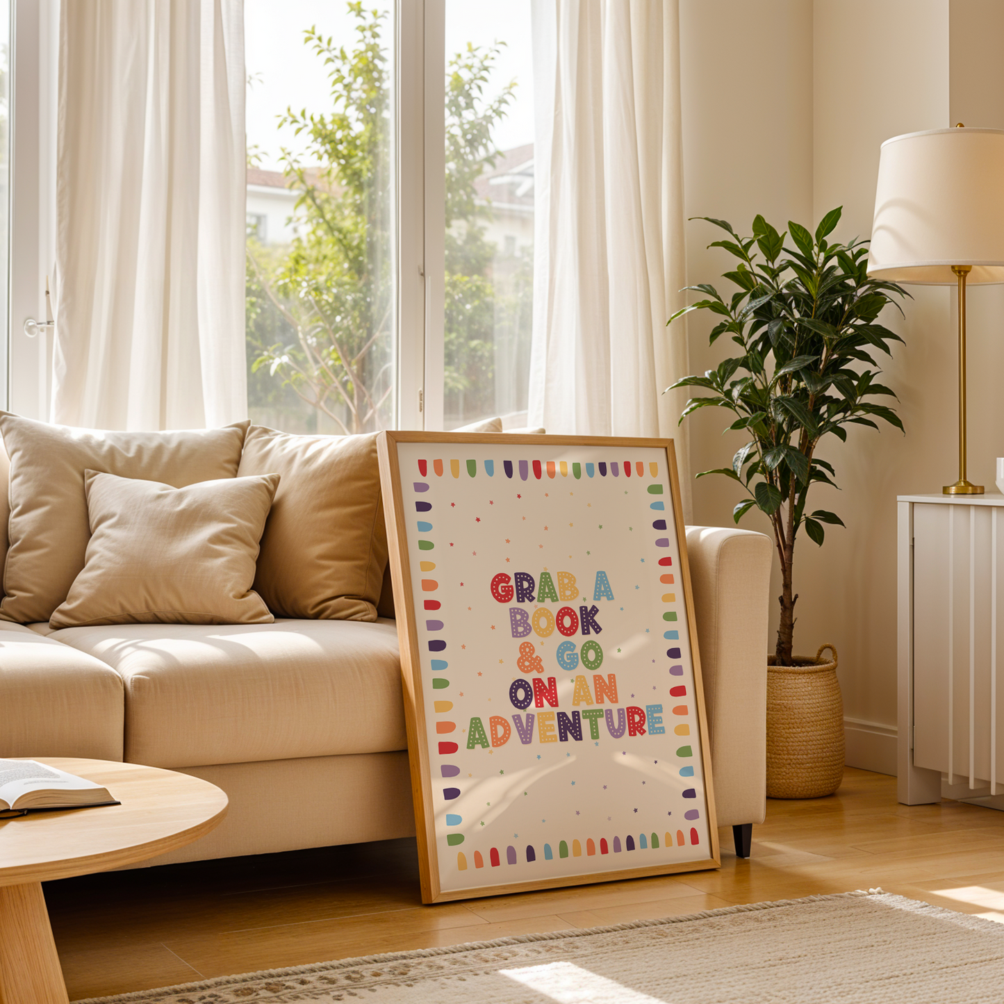 A framed children's book-themed art print leaning against a sofa on a light wood floor. The print features the phrase 'GRAB A BOOK & GO ON AN ADVENTURE' in colorful letters with a rainbow border.