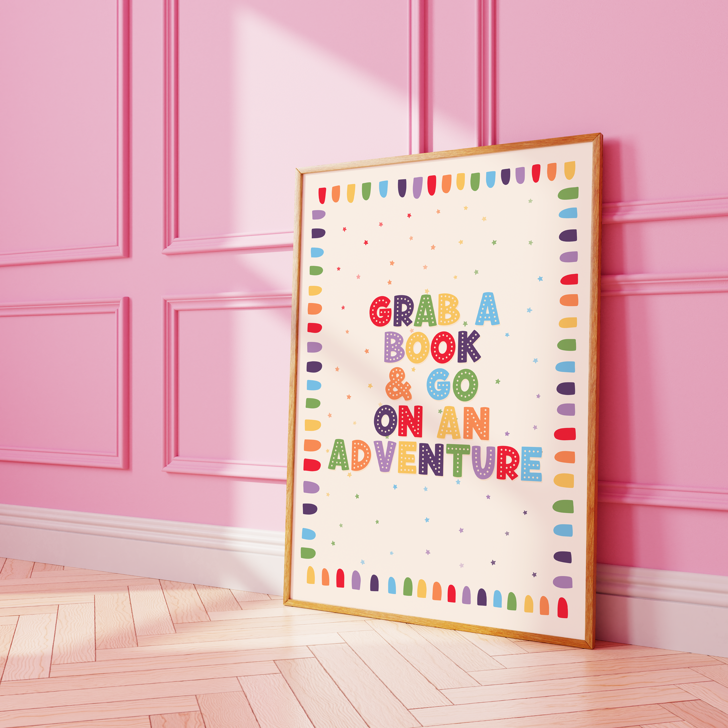 A framed children's book-themed art print leaning against a pink paneled wall on a light wood floor. The print features the phrase 'GRAB A BOOK & GO ON AN ADVENTURE' in colorful letters with a rainbow border.