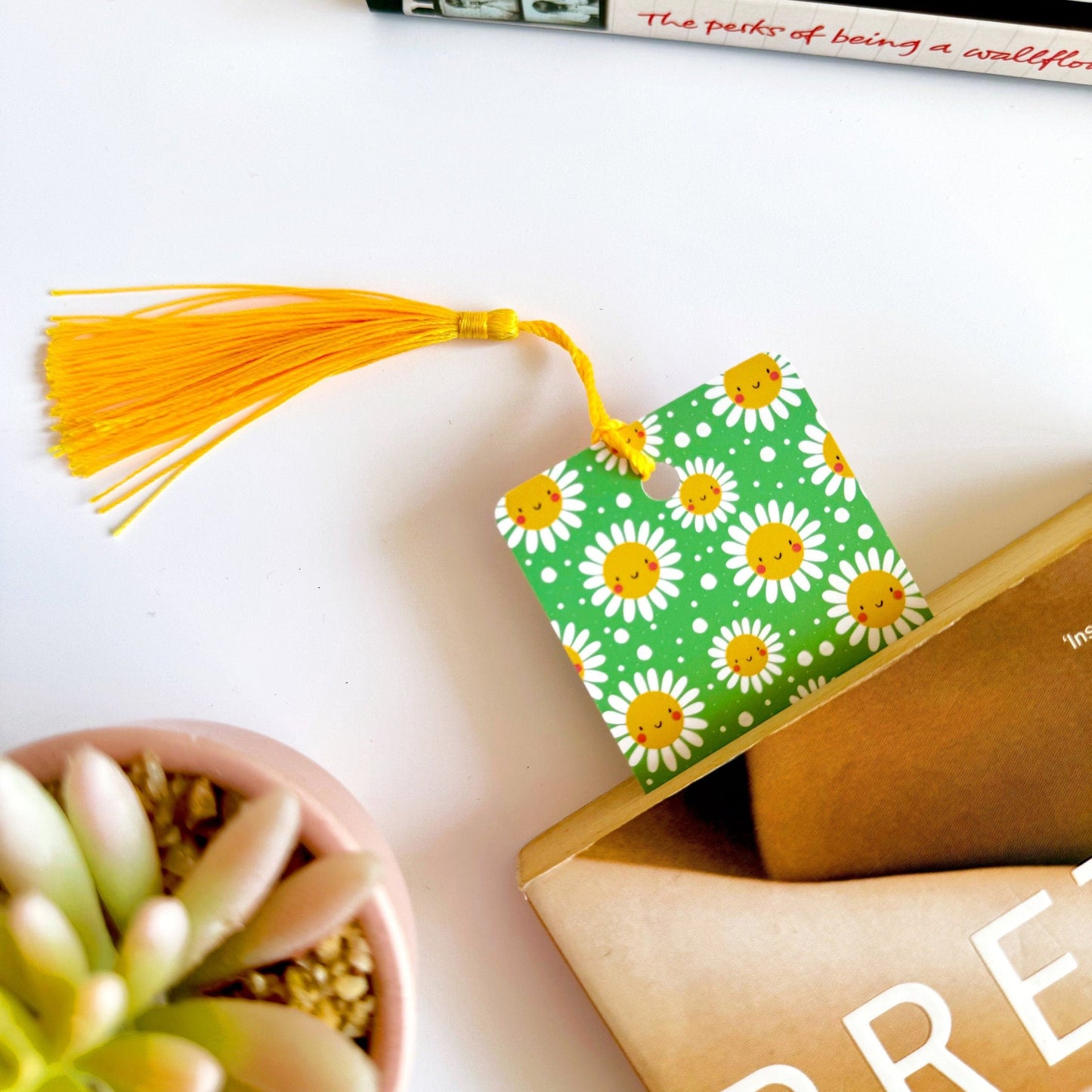 A picture of a green bookmark, with a coordinating yellow tassel, with a repeating pattern of white and yellow smiling flowers on it.