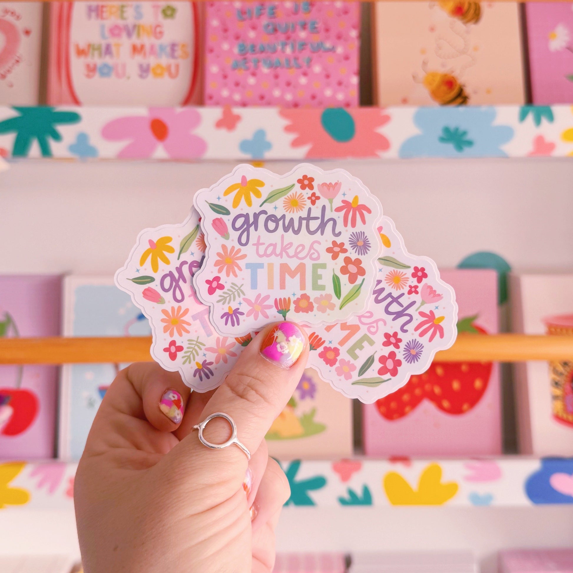 A holographic sticker with hand written text that reads 'growth takes time', which is decorated with lots of floral doodles.
