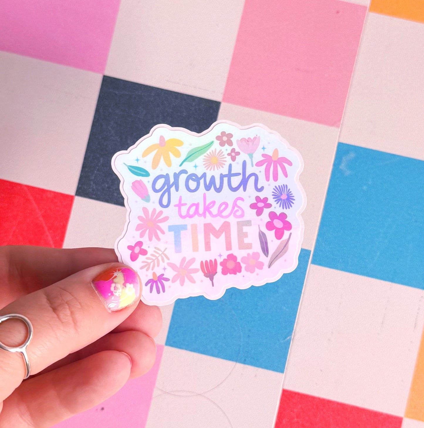 A holographic sticker with hand written text that reads 'growth takes time', which is decorated with lots of floral doodles.