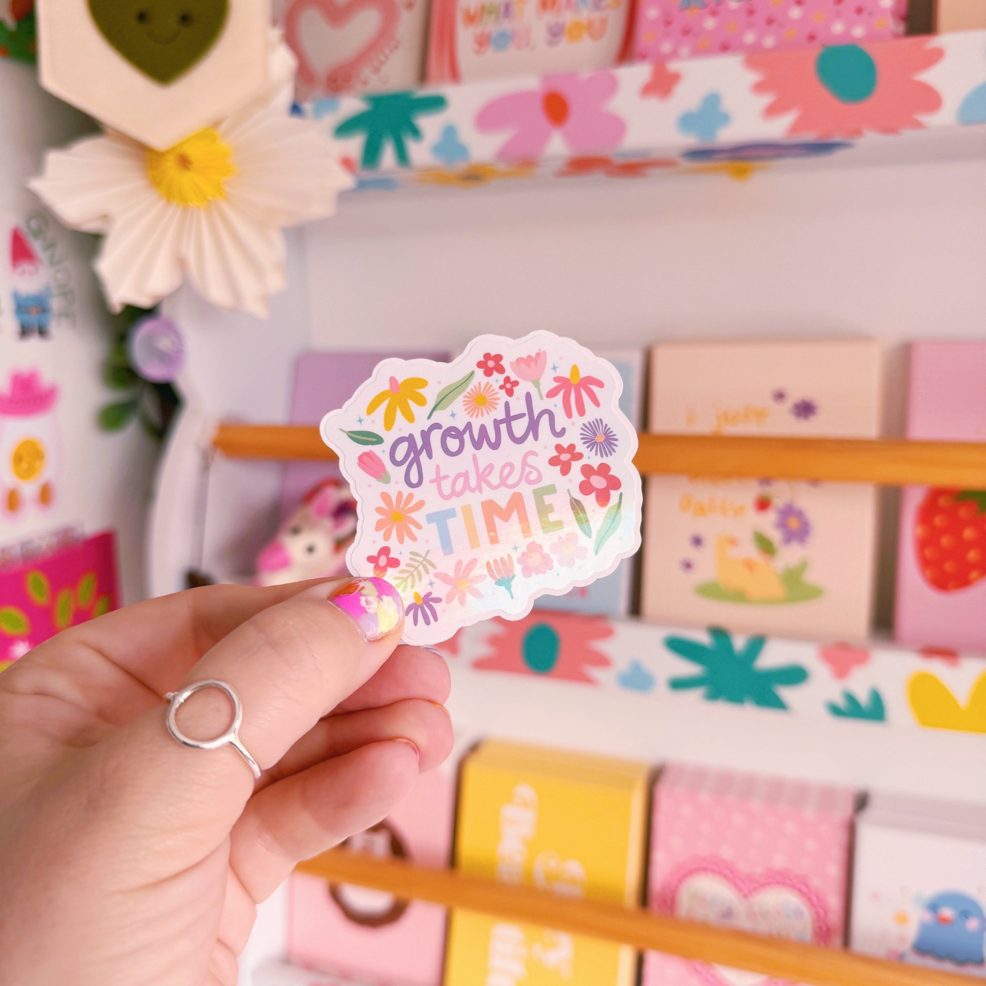 A holographic sticker with hand written text that reads 'growth takes time', which is decorated with lots of floral doodles.