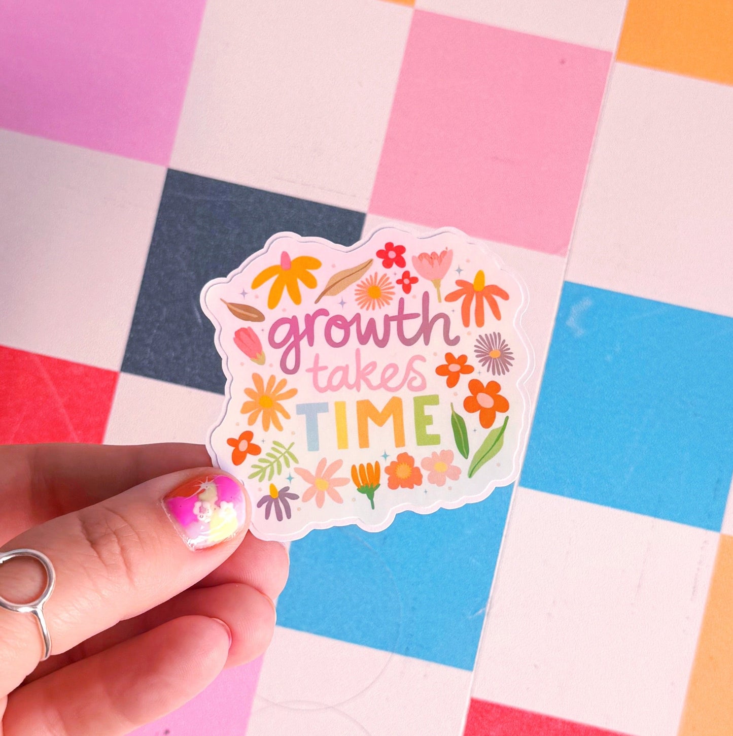 A holographic sticker with hand written text that reads 'growth takes time', which is decorated with lots of floral doodles.