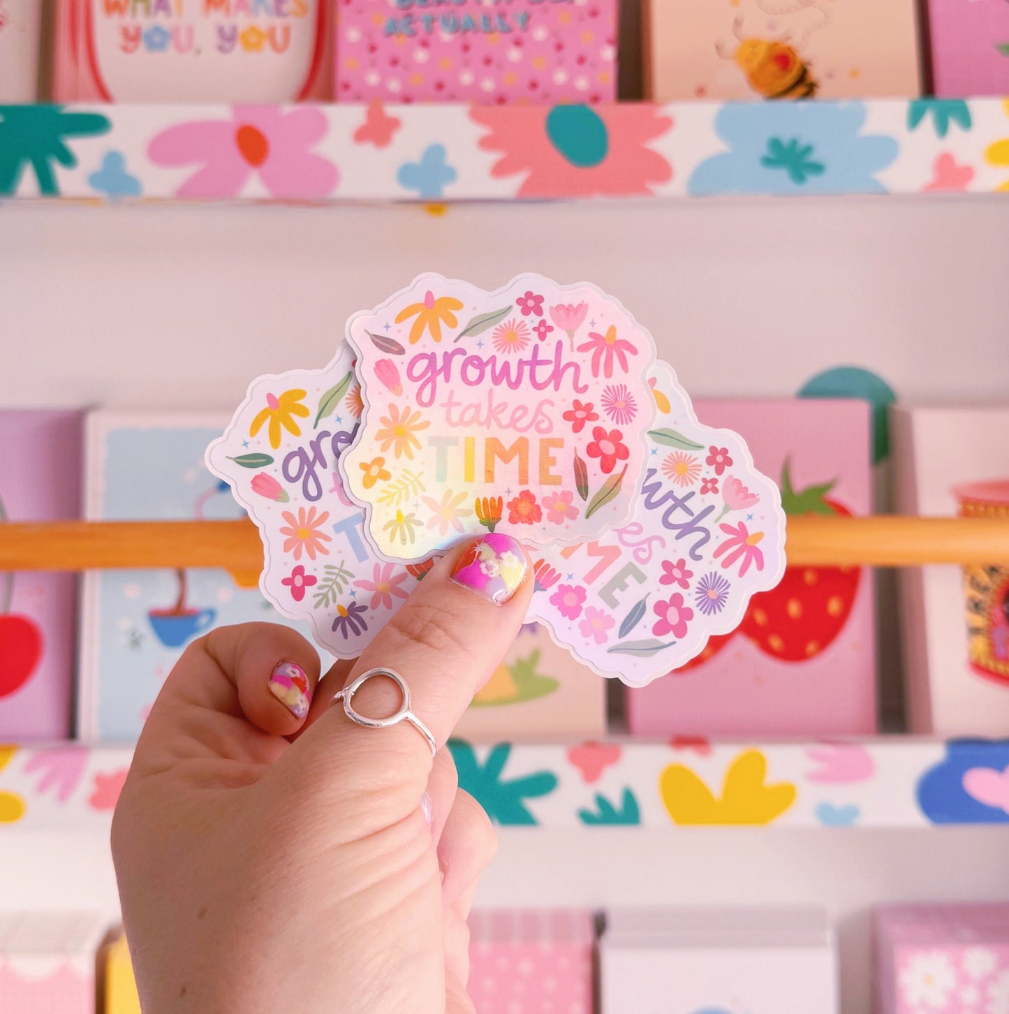 A holographic sticker with hand written text that reads 'growth takes time', which is decorated with lots of floral doodles.