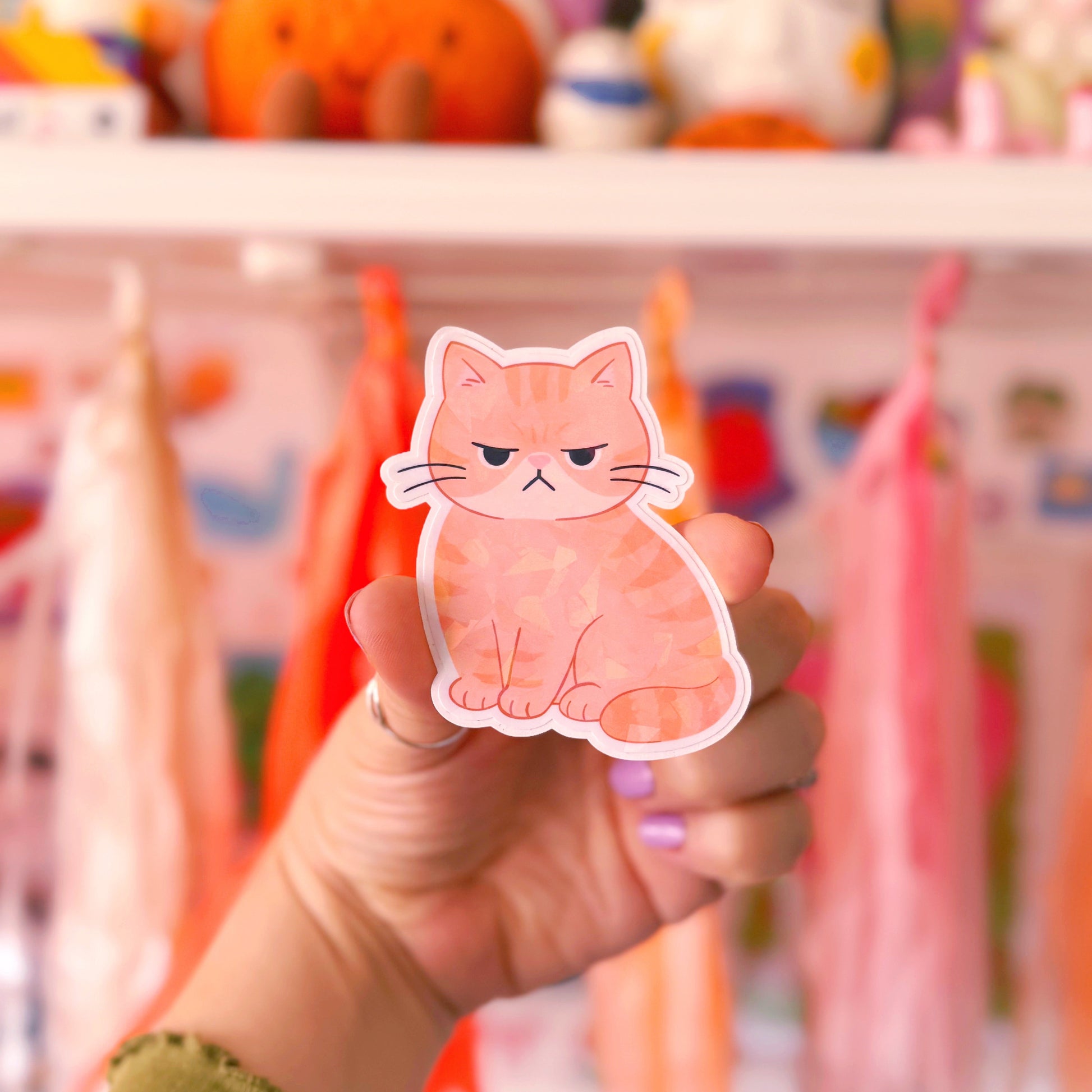 A hand holding a die-cut sticker of a ginger cat with a grumpy expression, set against a blurred background of colourful shelves and plush toys.