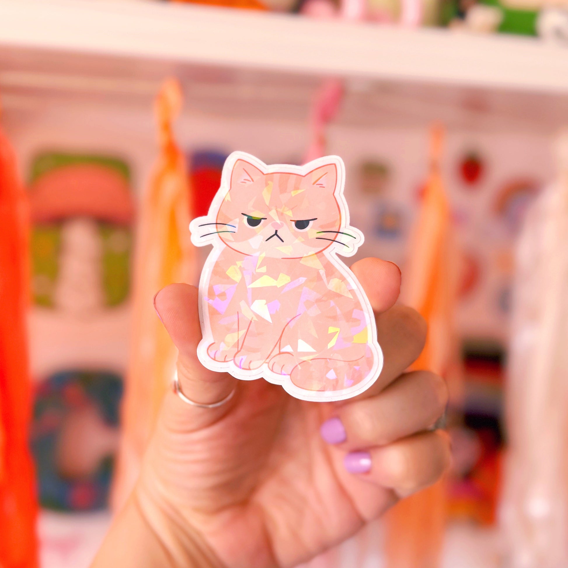A hand holding a die-cut sticker of a ginger cat with a grumpy expression, set against a blurred background of colourful shelves and plush toys.