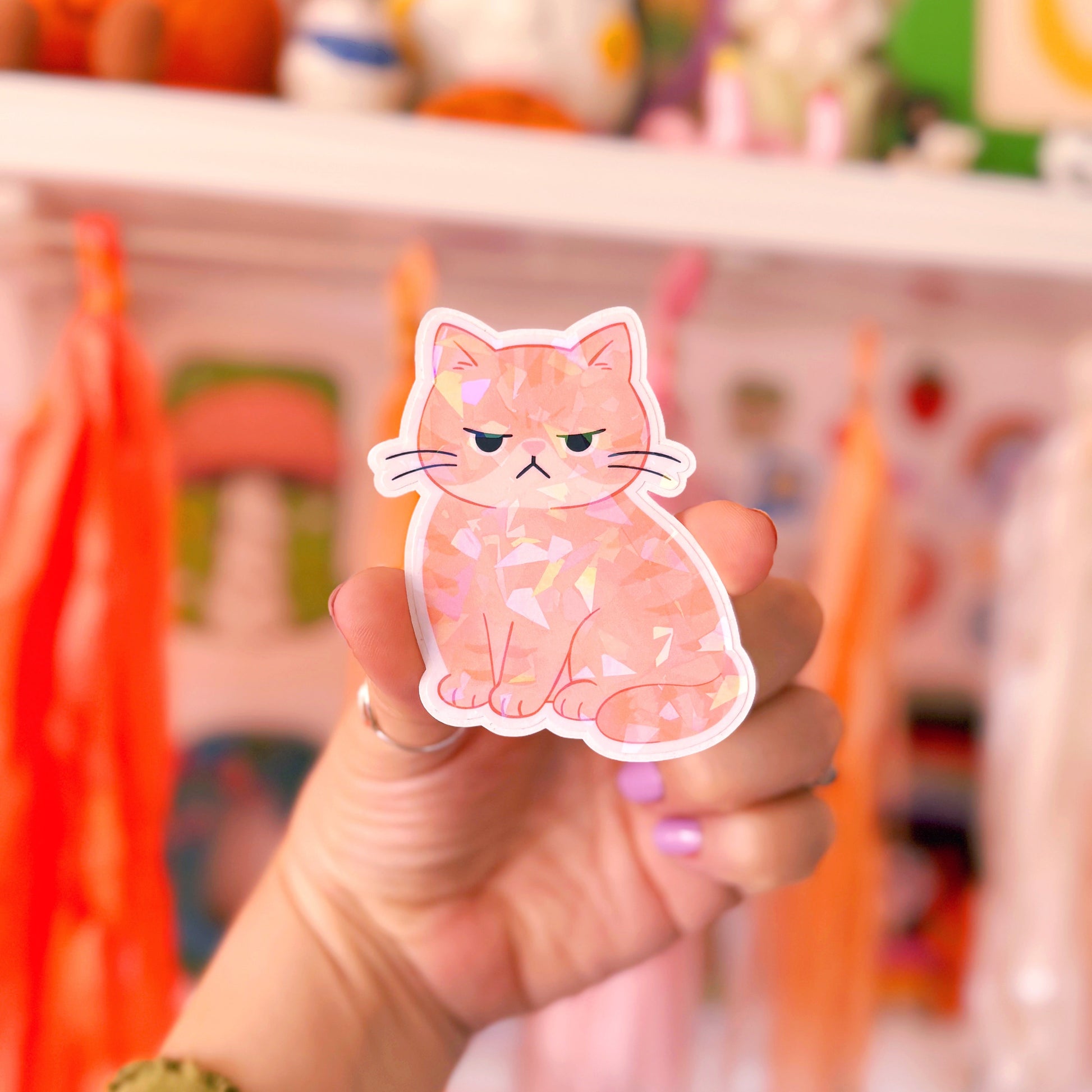 A hand holding a die-cut sticker of a ginger cat with a grumpy expression, set against a blurred background of colourful shelves and plush toys.