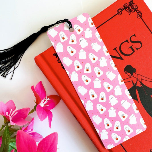 A picture of a pink bookmark, with a coordinating black tassel, with a repeating pattern of white cute ghosts all over.