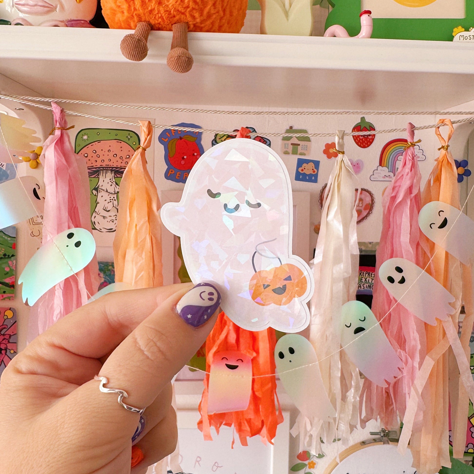 Hand holding a ghost-shaped sticker with a colourful background featuring toys and decorations.