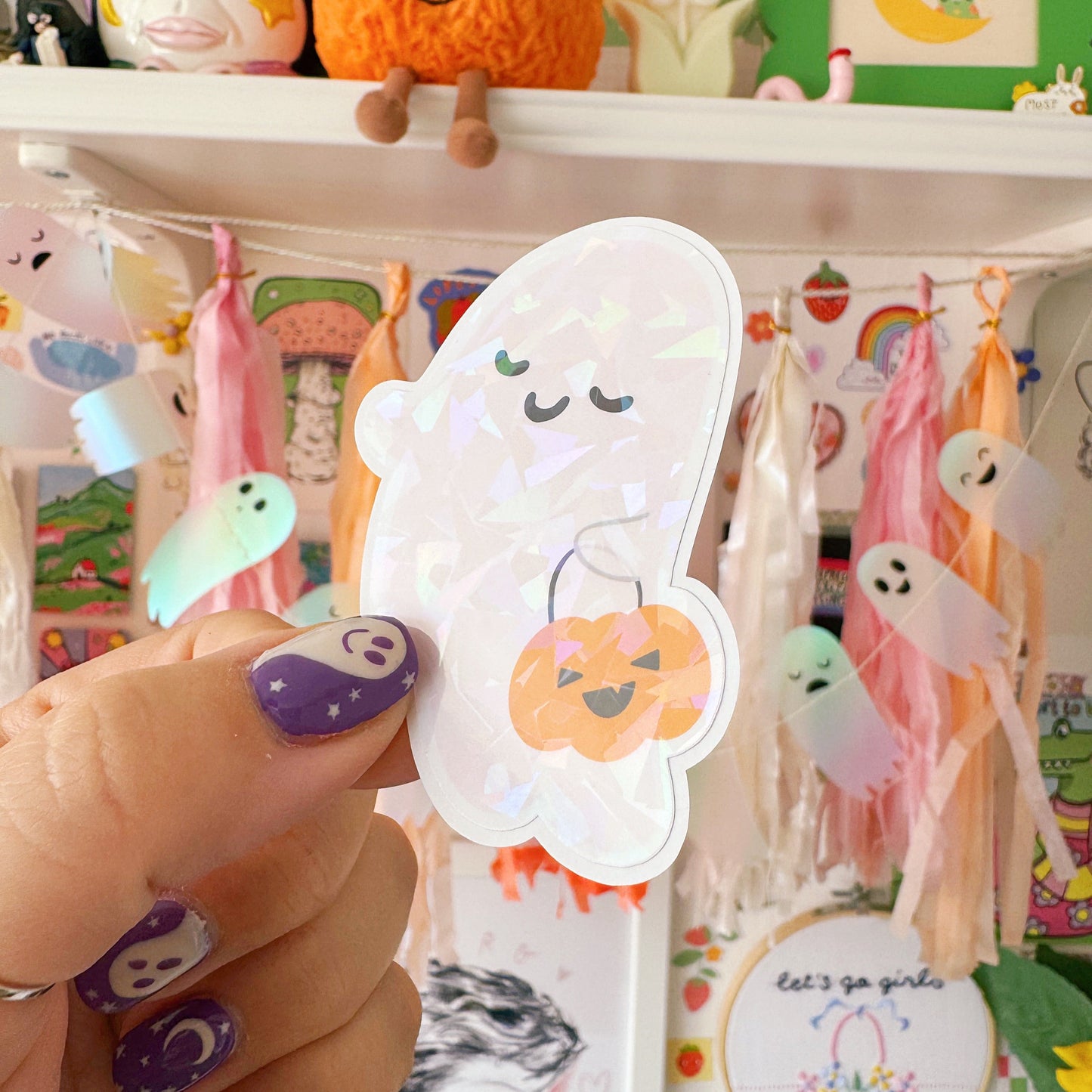 Hand holding a cute ghost-shaped Halloween sticker with a colorful background of stationery items.