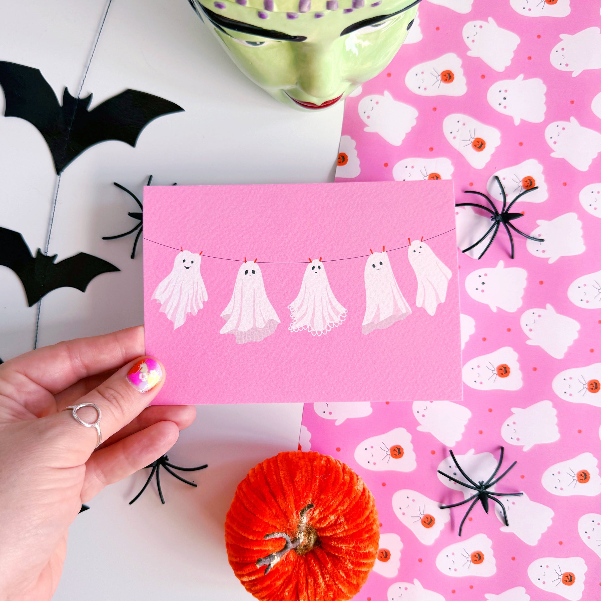 A halloween themed postcard of 5 ghosts hanging on a washing line, on a pink background.