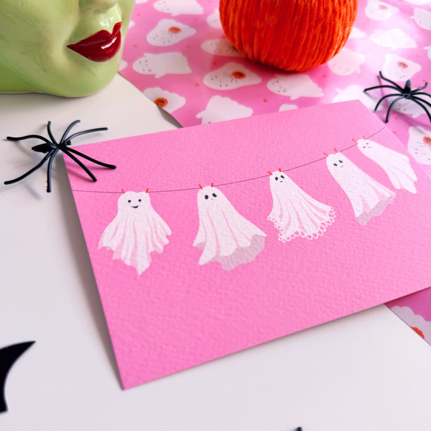 A halloween themed postcard of 5 ghosts hanging on a washing line, on a pink background.