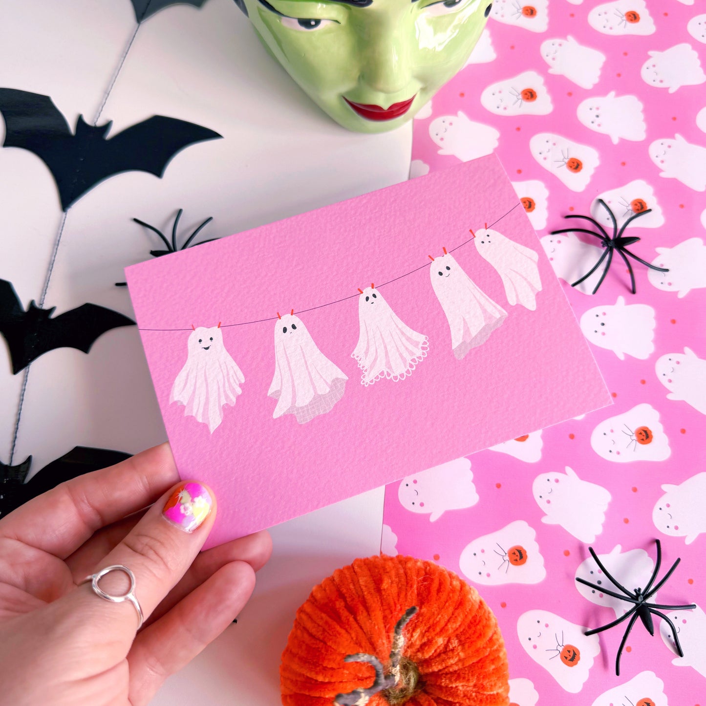 A halloween themed postcard of 5 ghosts hanging on a washing line, on a pink background.
