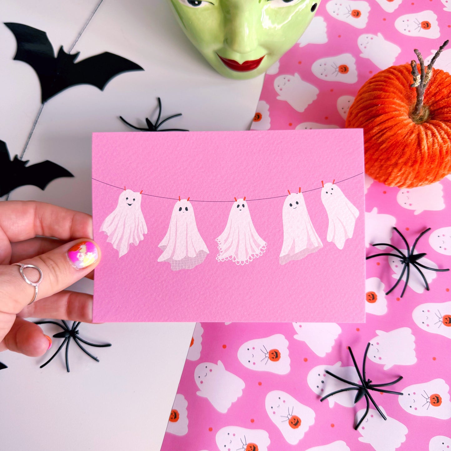 A halloween themed postcard of 5 ghosts hanging on a washing line, on a pink background.