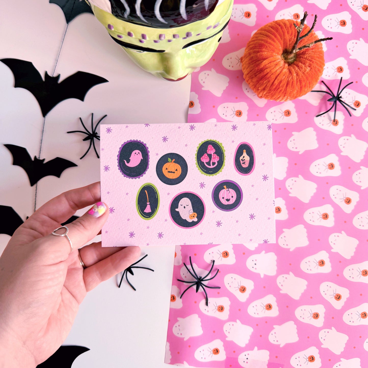 A landscape postcard with 7 illustrated frames, inside of which are spooky cute Halloween illustrations. The background of the postcard is pale purple with purple starbursts.