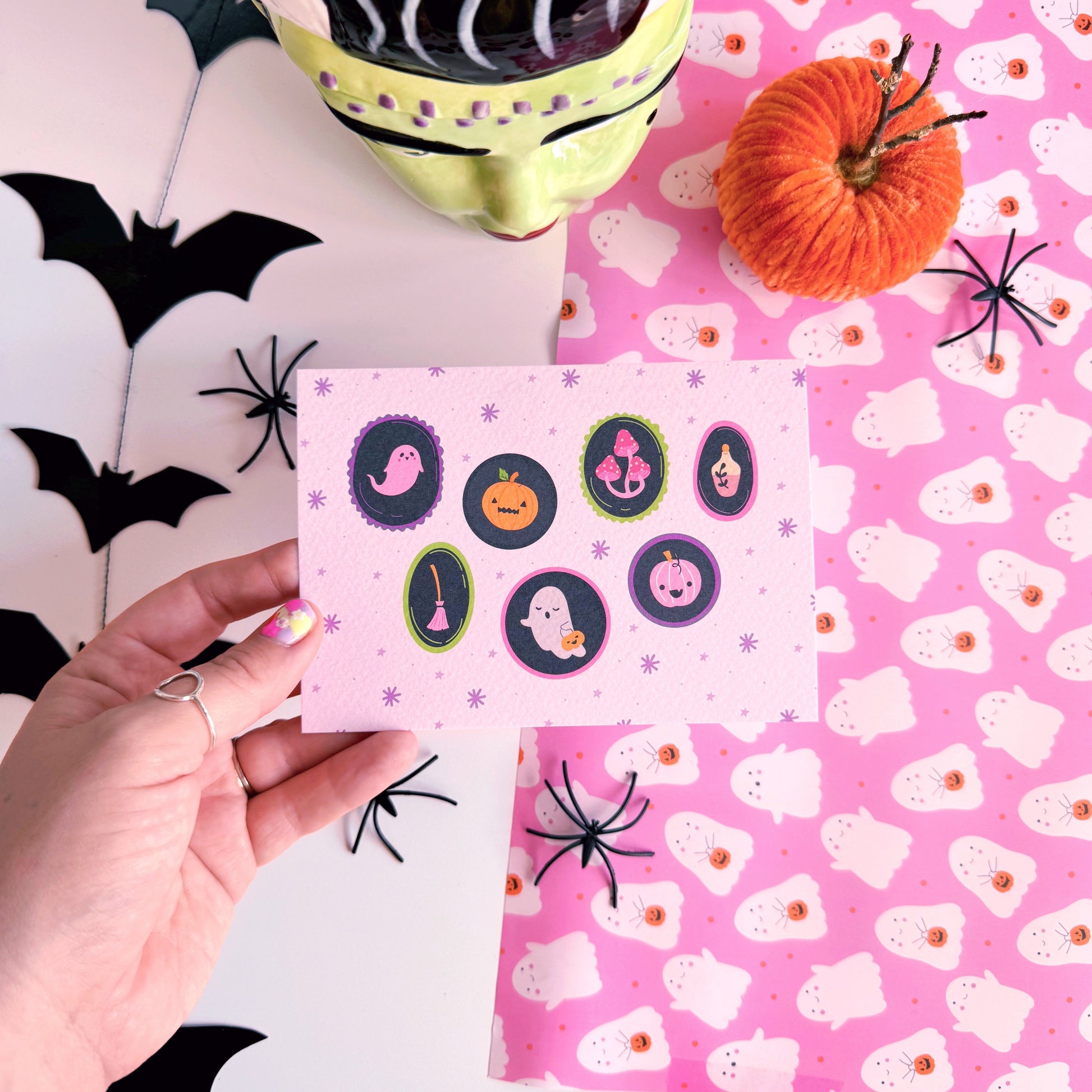 A landscape postcard with 7 illustrated frames, inside of which are spooky cute Halloween illustrations. The background of the postcard is pale purple with purple starbursts.