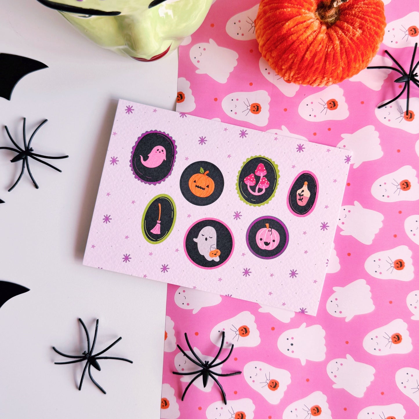 A landscape postcard with 7 illustrated frames, inside of which are spooky cute Halloween illustrations. The background of the postcard is pale purple with purple starbursts.