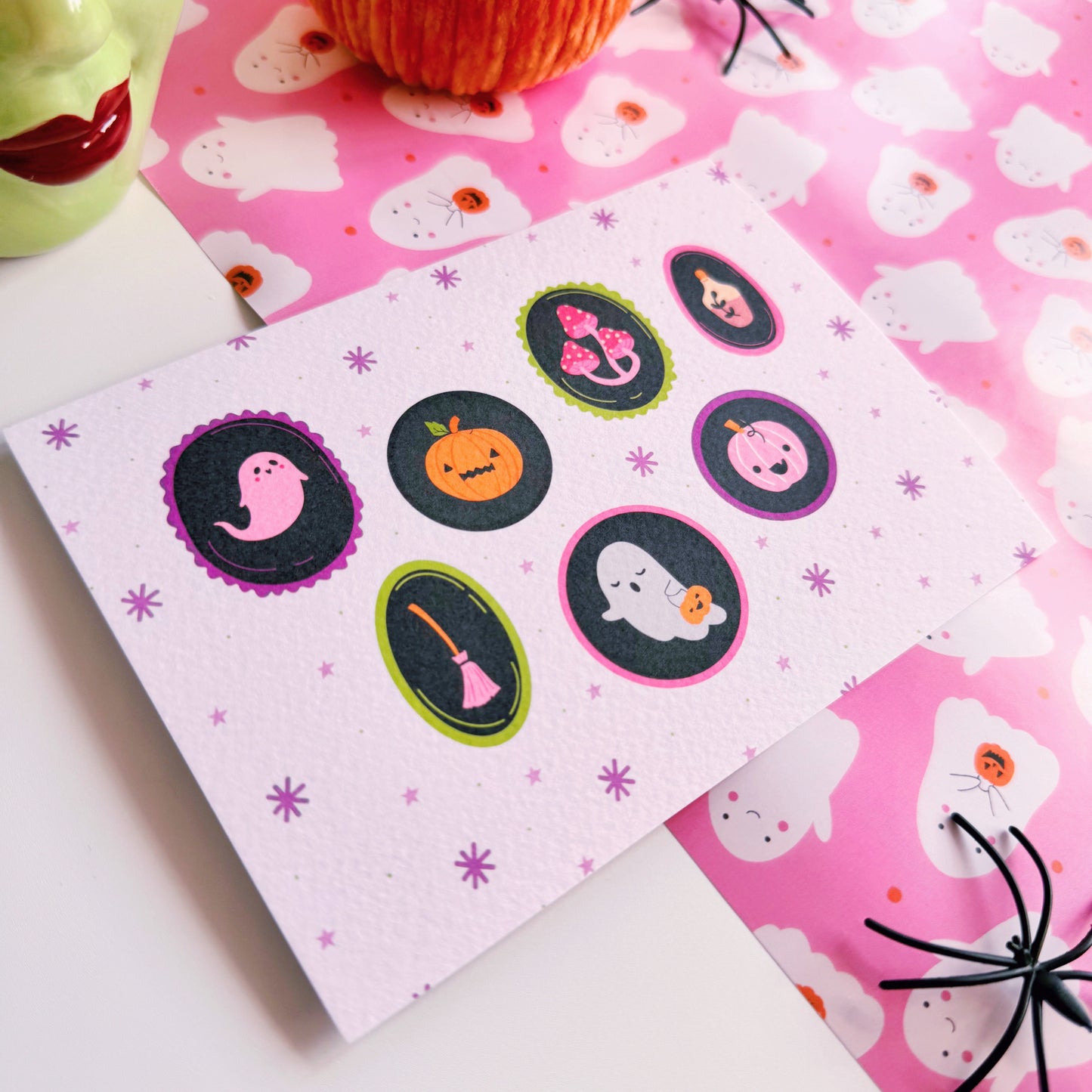 A landscape postcard with 7 illustrated frames, inside of which are spooky cute Halloween illustrations. The background of the postcard is pale purple with purple starbursts.