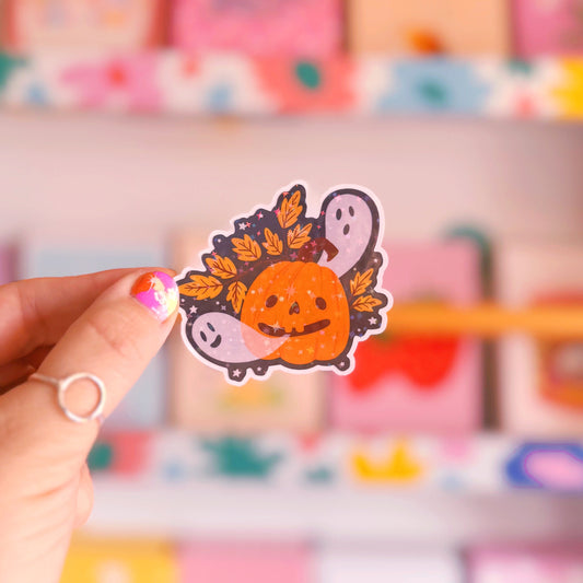 A holographic sticker with an illustration of a smiling pumpkin, with ghosts coming out of his mouth.