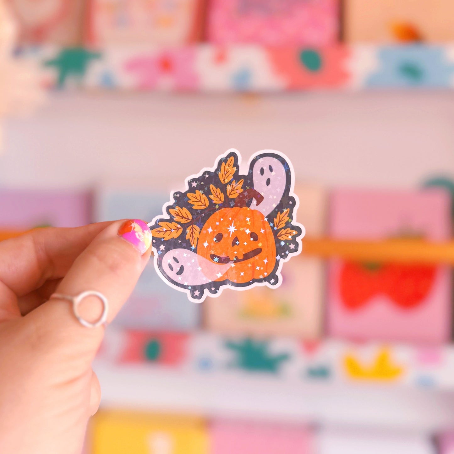 A holographic sticker with an illustration of a smiling pumpkin, with ghosts coming out of his mouth.