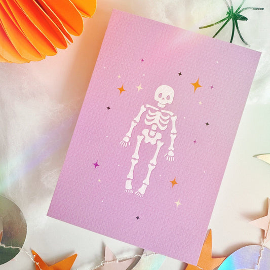 A halloween themed postcard of a cute spooky skeleton surrounded by stars, on a purple background.